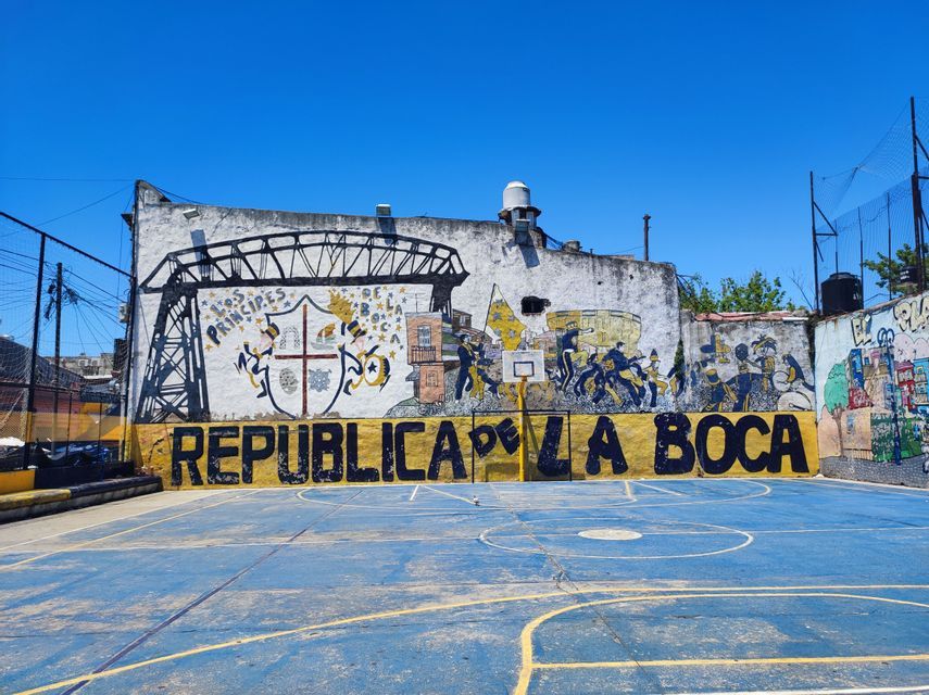 Un campo da basket blu vuoto si trova di fronte a un muro con un grande murale che recita 'Republica de La Boca' sotto un cielo limpido.