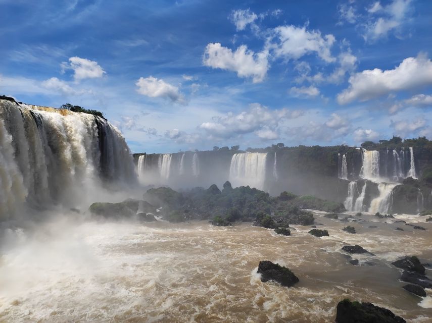 Una vista panorámica de potentes cascadas que caen en un río marrón turbulento, con niebla ascendiendo sobre un telón de fondo de árboles verdes bajo un cielo azul nublado.