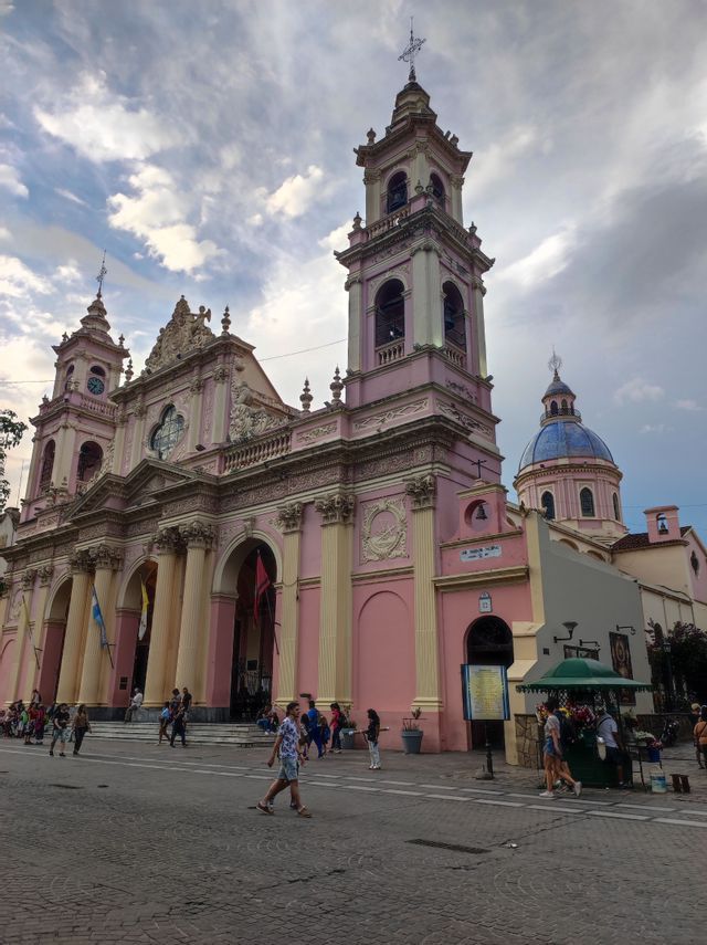 Una cattedrale rosa riccamente decorata, con colonne color crema e due campanili gemelli, si erge su una piazza acciottolata mentre le persone passeggiano.