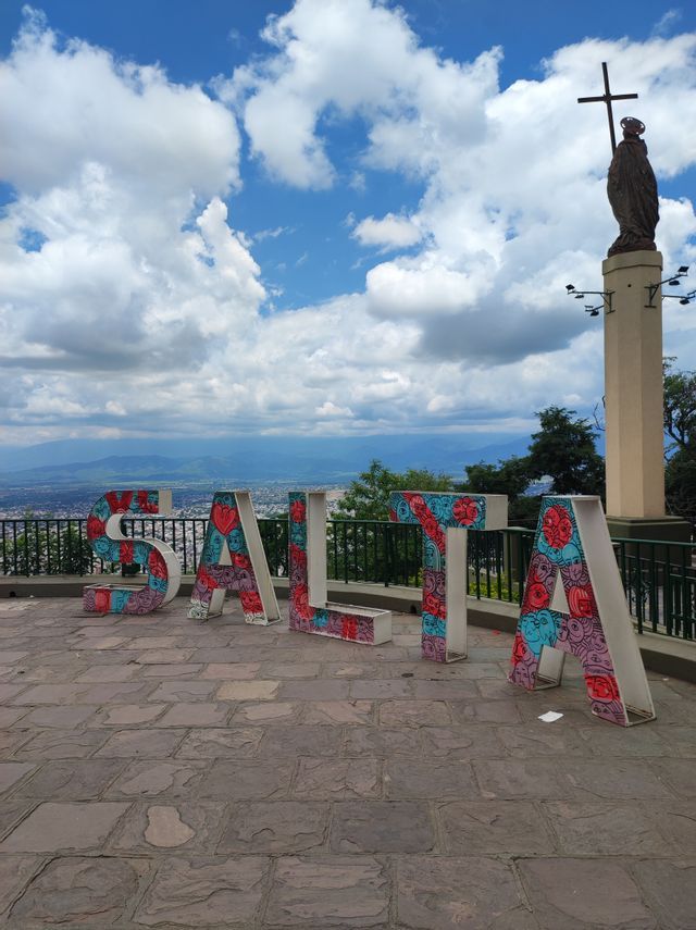 Un'insegna colorata che forma la parola SALTA su un punto panoramico in pietra, con una statua su un pilastro che domina un paesaggio urbano sotto un cielo nuvoloso.