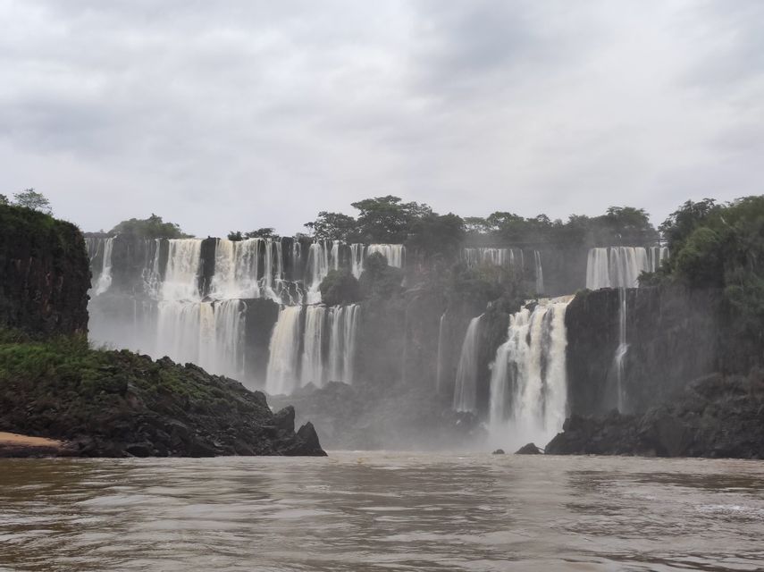 Una cascada amplia y de varios niveles fluye sobre acantilados rocosos cubiertos de árboles, cayendo en un río bajo un cielo nublado.