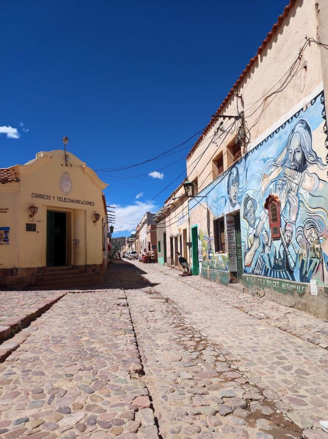 Una strada acciottolata passa tra un edificio giallo e un altro con un grande murale colorato sotto un cielo azzurro e limpido.