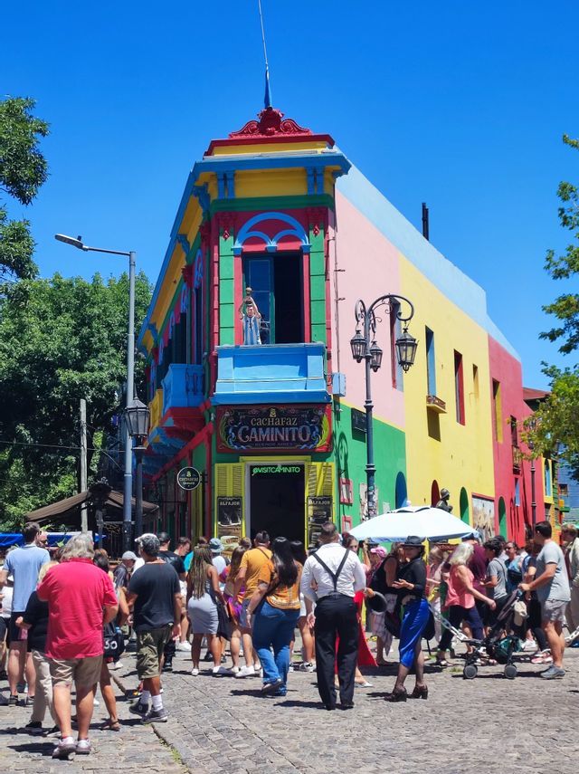 Una folla di persone su una strada di ciottoli davanti a un edificio angolare multicolore in una giornata di sole.