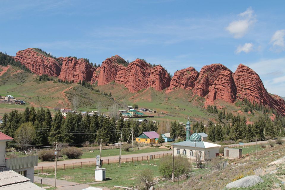 Un petit village avec une mosquée est niché au pied de grandes formations rocheuses rouges superposées, sur une colline verdoyante sous un ciel bleu.