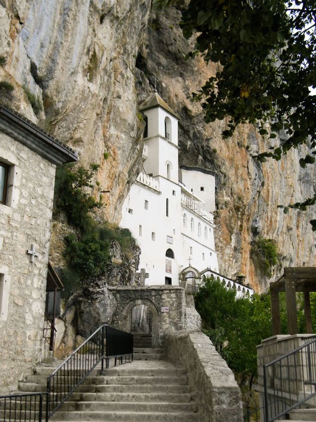 Ein weißes, mehrstöckiges Kloster mit Glockenturm ist in eine steile Felswand gebaut, mit einer Steintreppe im Vordergrund.