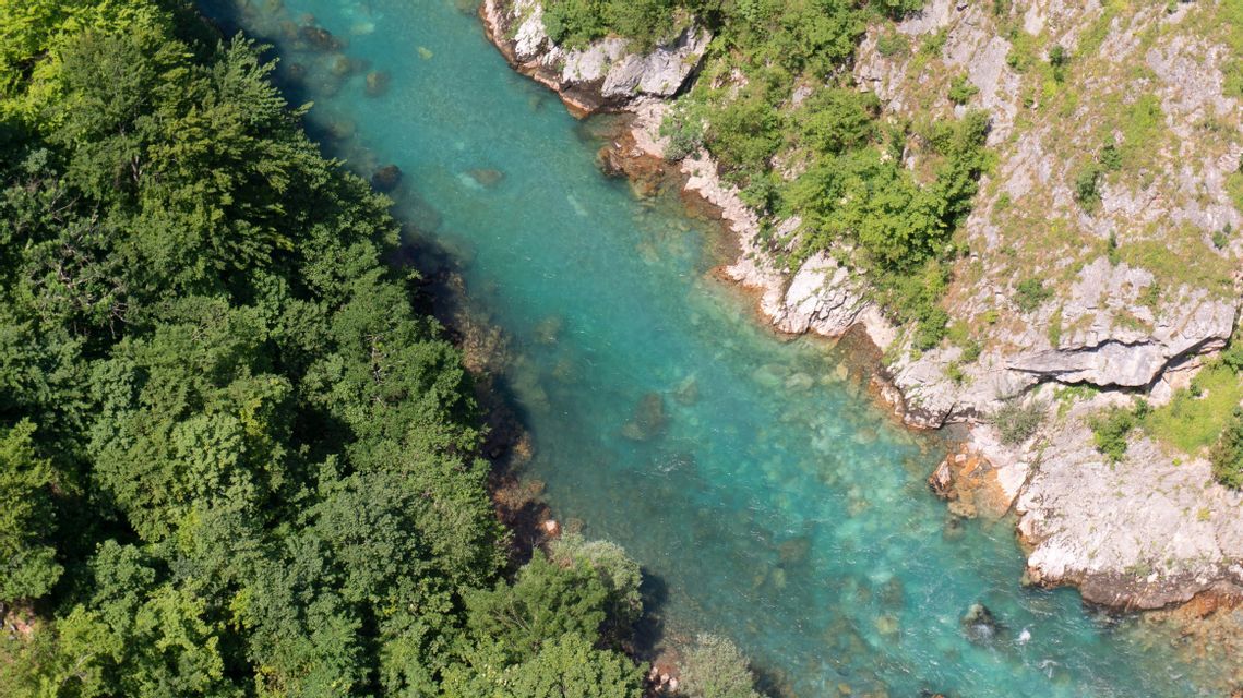 Eine Luftaufnahme zeigt einen türkisfarbenen Fluss, der zwischen einem dichten grünen Wald und einer steilen, felsigen Canyonwand verläuft.
