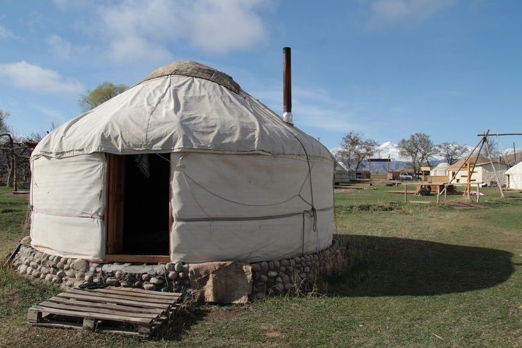 Une yourte blanche traditionnelle avec une base en pierre se trouve dans un camp herbeux, avec des montagnes enneigées visibles en arrière-plan.
