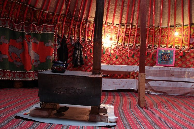 L'intérieur d'une yourte traditionnelle avec un poêle à bois central, une théière bleue et des textiles à motifs rouges recouvrant les murs et le sol.