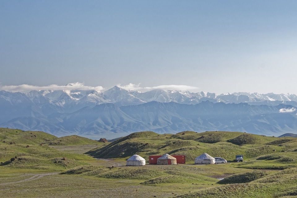 Un camp de yourtes avec des véhicules nichés dans des collines verdoyantes devant une chaîne de montagnes enneigée et lointaine.