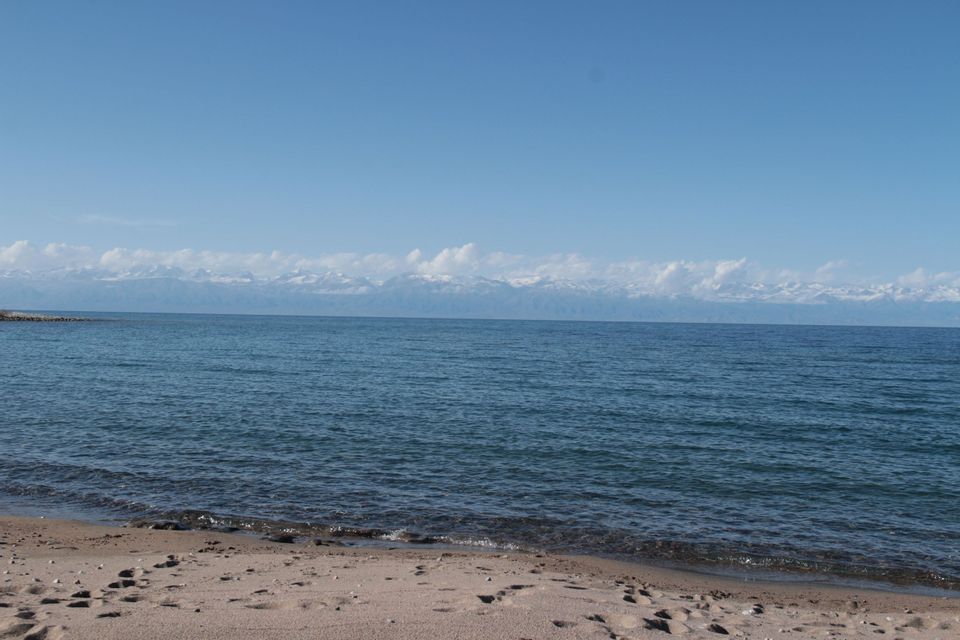 Une plage de sable rencontre l'eau bleue calme d'un lac, avec des montagnes enneigées visibles au loin sous un ciel clair.