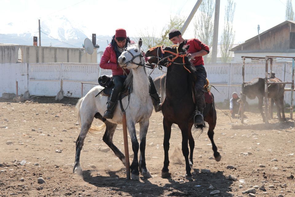 Deux hommes à cheval, l'un sur un cheval blanc et l'autre sur un cheval brun, se font face dans un enclos poussiéreux avec des montagnes en arrière-plan.