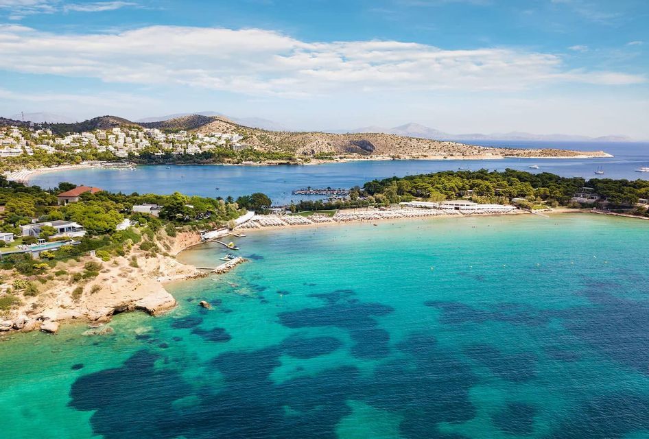 Vista aerea di una costa con acqua turchese cristallina, spiagge sabbiose e una cittadina con edifici bianchi su una collina verdeggiante.