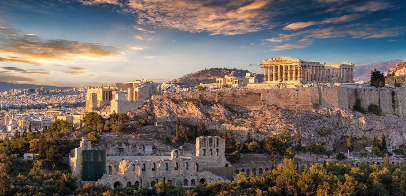 Eine historische Akropolis mit antiken Ruinen thront auf einem felsigen Hügel und blickt auf eine weitläufige Stadt unter einem farbenprächtigen Sonnenuntergangshimmel.