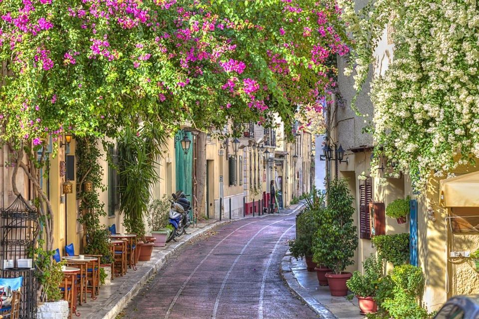 Una strada stretta e sinuosa fiancheggiata da caffè e ricoperta da cascate di viti fiorite rosa e bianche.