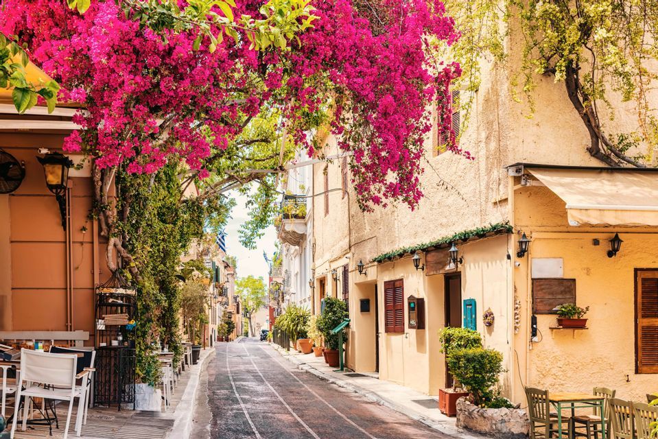 Una via stretta e soleggiata è fiancheggiata da tavolini di caffè all'aperto e incorniciata da un arco di vivaci bougainvillee rosa.
