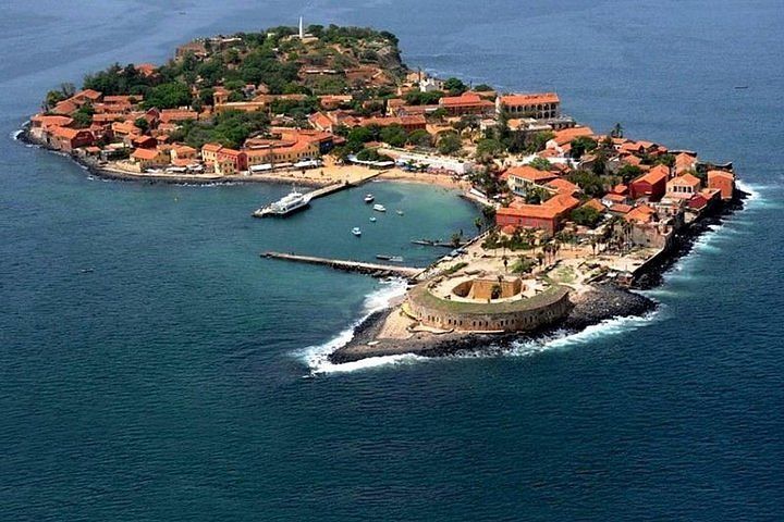 An aerial view of a small island with red-roofed buildings, a harbor with boats, and a stone fort in the blue sea.