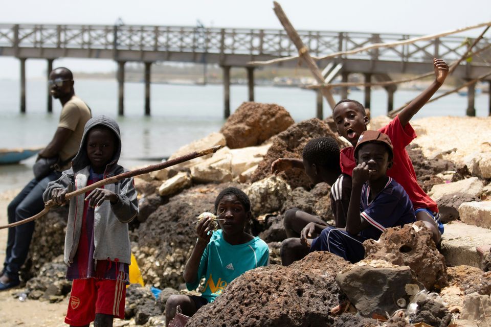 A group of children sitting and playing on a rocky shore, with a wooden pier spanning the water in the background.