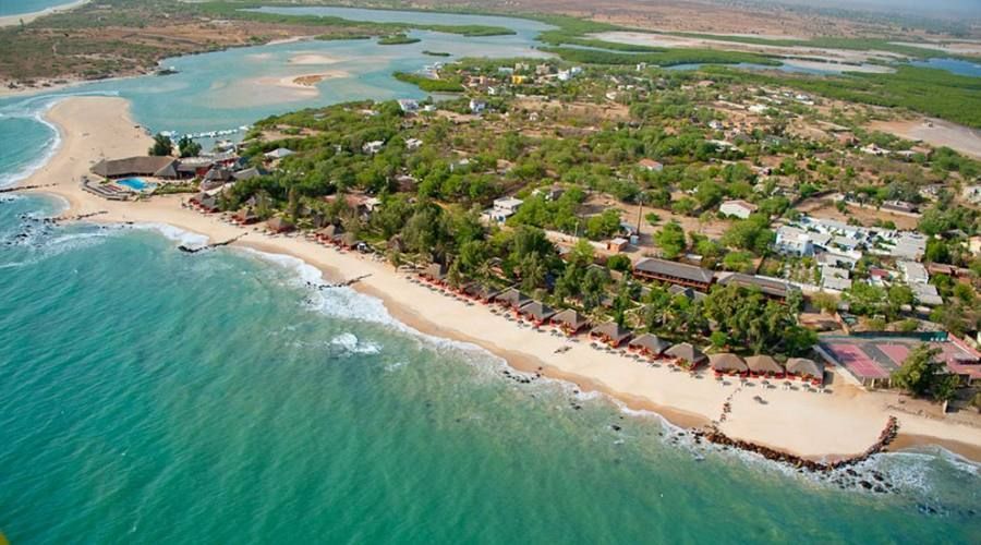 An aerial view of a coastal resort village with thatched-roof huts along a sandy beach and turquoise ocean.