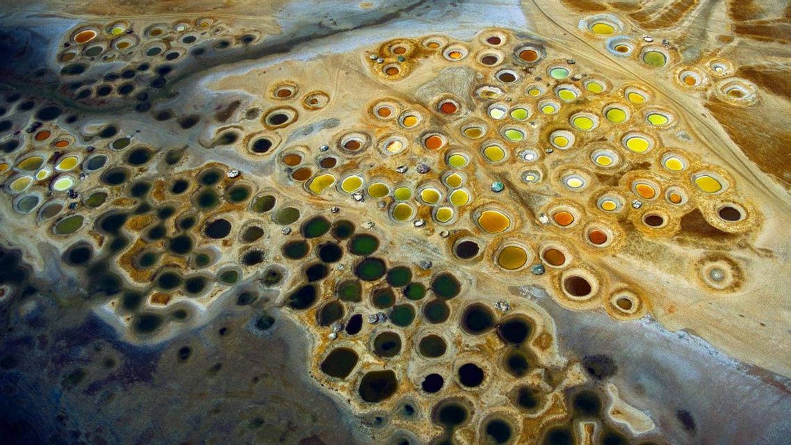 Una vista aerea di un paesaggio ricco di numerose pozze d'acqua circolari nelle tonalità del verde, giallo e arancione.