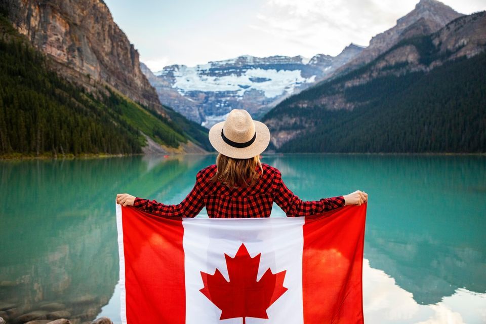 Une personne de dos, vêtue d'une chemise à carreaux et d'un chapeau, tient un drapeau canadien tout en admirant un lac alpin turquoise et des montagnes.