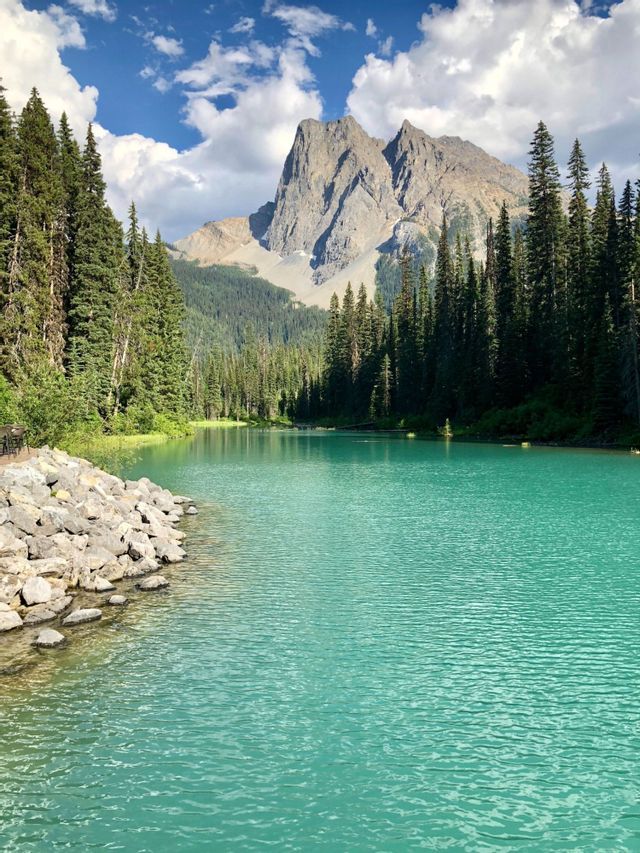 Un lac turquoise avec une rive rocheuse est entouré d'une forêt de pins, avec un sommet montagneux s'élevant à l'arrière-plan sous un ciel nuageux.