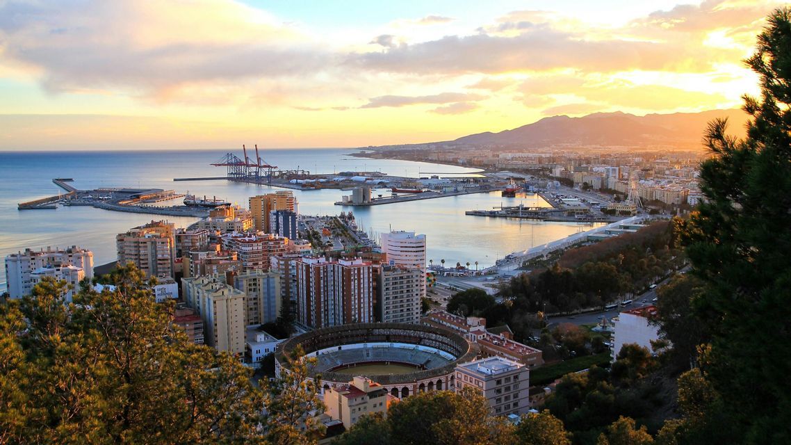 An elevated view of a coastal city with a large port and a circular bullring, all under a golden sunset sky.