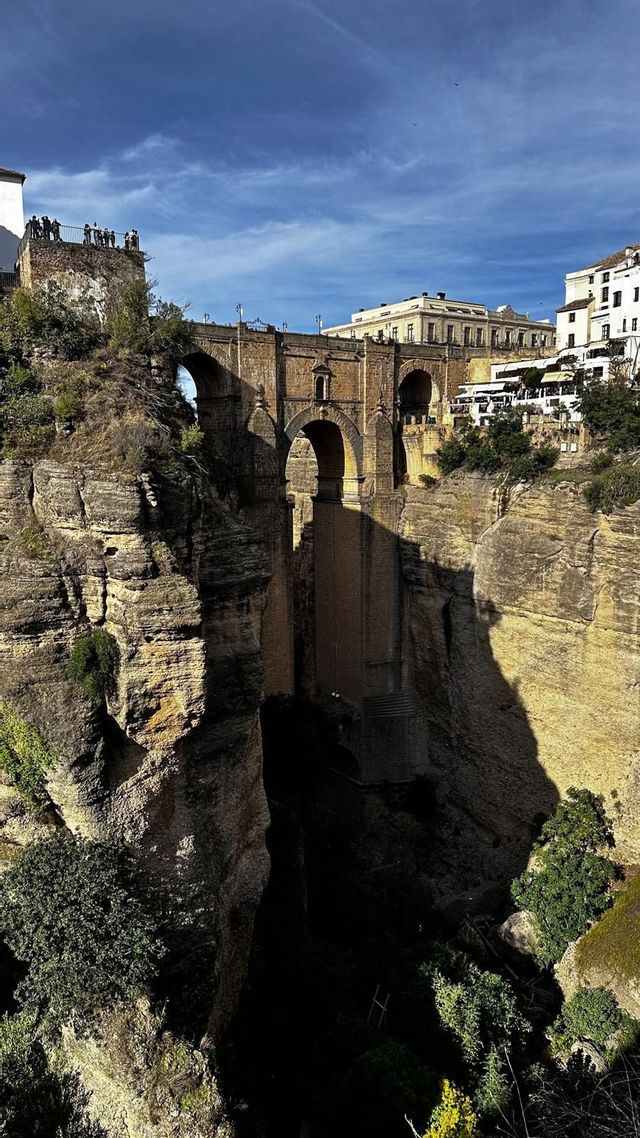 An arched stone bridge spans a deep rocky gorge, with white buildings on the cliffs and a WeRoad group trip at a viewpoint.