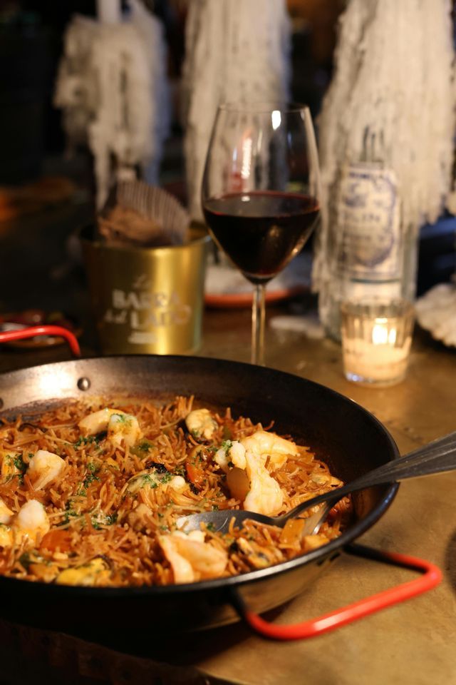 A pan of Spanish fideuà with seafood sits on a table next to a glass of red wine and a lit candle.