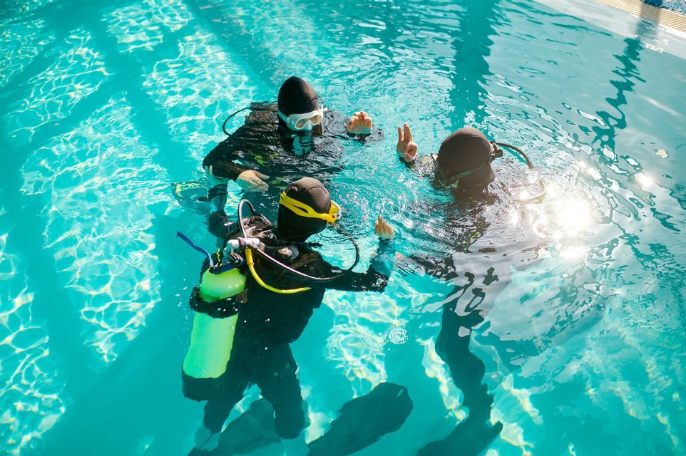 Un voyage en groupe WeRoad de trois personnes, équipées de matériel de plongée, apprenant des techniques de plongée dans une piscine ensoleillée.