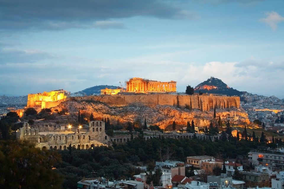 Una antigua ciudadela, brillantemente iluminada con luces doradas, se asienta sobre una colina rocosa con vistas a un paisaje urbano al anochecer.