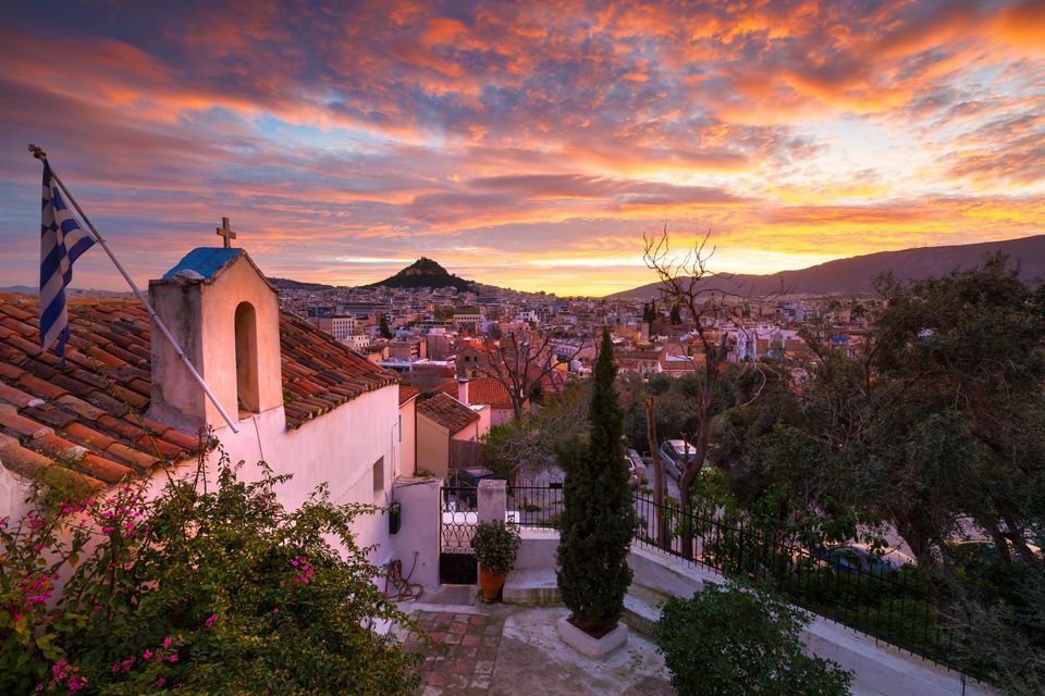 Una chiesa imbiancata con una bandiera greca sul tetto domina un paesaggio urbano con una collina in lontananza sotto un cielo al tramonto dai colori vivaci.