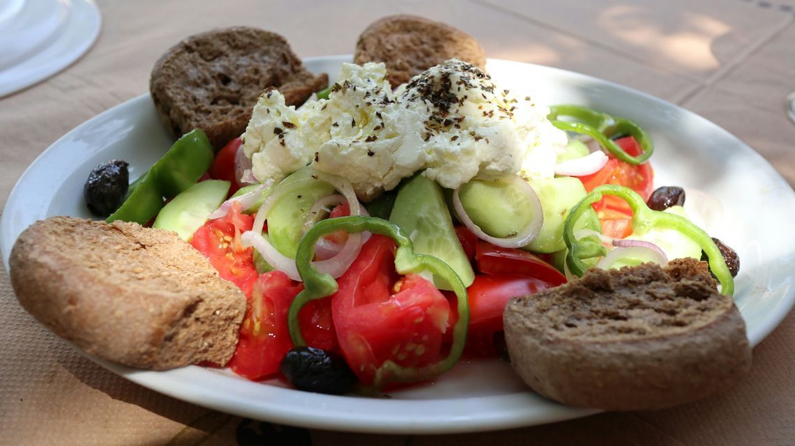 Primo piano di un'insalata greca con pomodori, cetrioli, peperoni, formaggio feta e olive, servita con pane scuro su un piatto bianco.