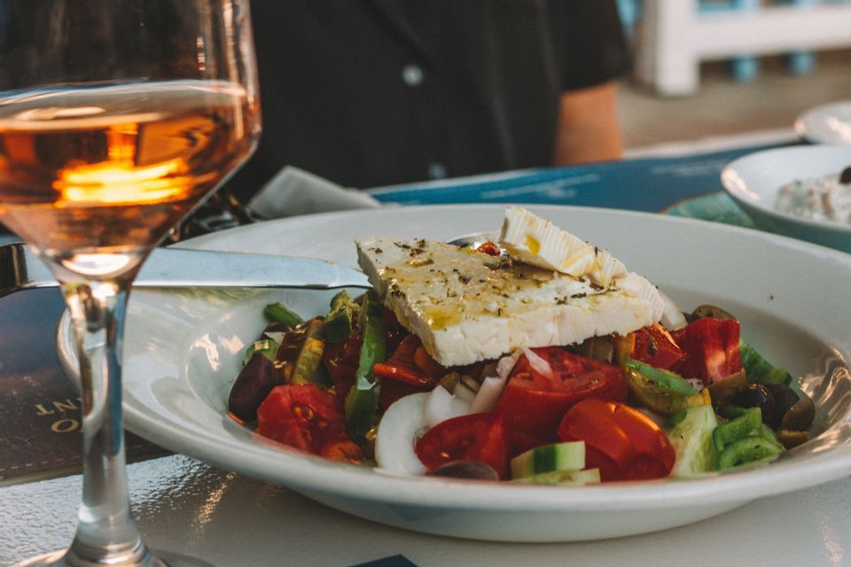 Una ciotola bianca di insalata greca con formaggio feta, accanto a un bicchiere di vino rosato su un tavolo.
