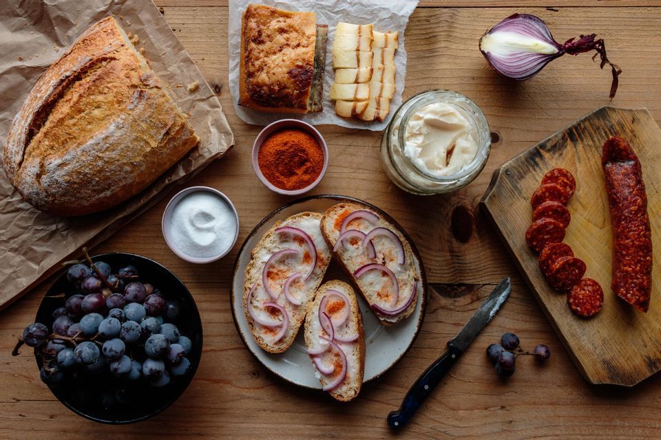Una vista dall'alto di una tavola rustica imbandita con pane, lardo, cipolla rossa, salame a fette e uva.