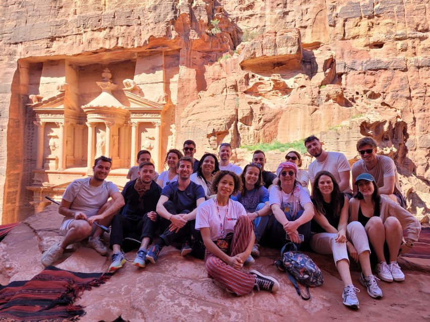 Un groupe WeRoad en voyage pose pour une photo de groupe sur une corniche rocheuse, devant une façade ancienne sculptée dans une falaise rouge.