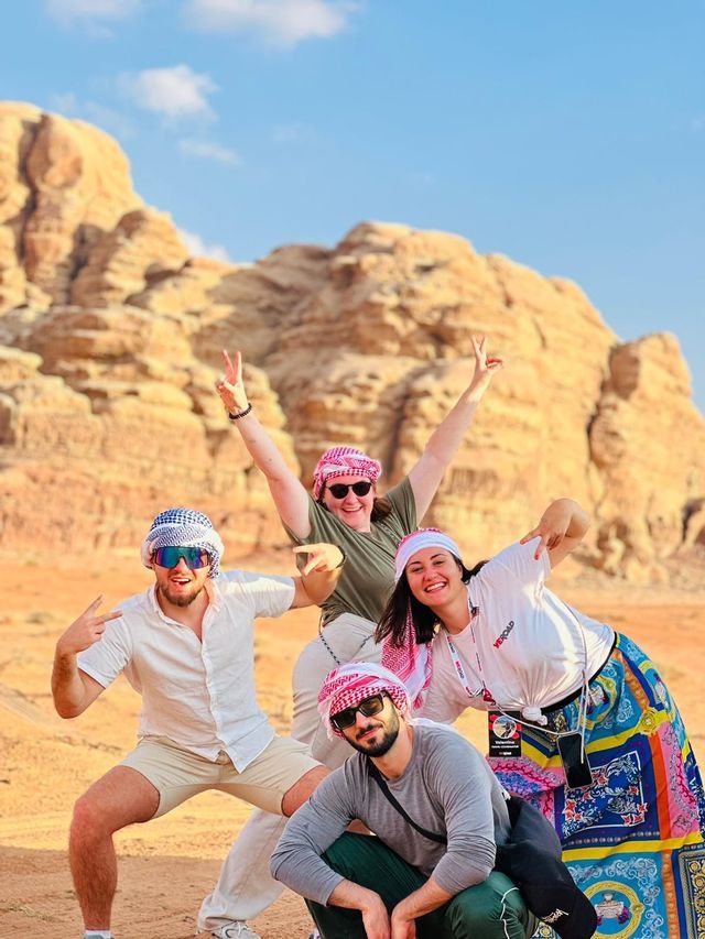 Un voyage de groupe WeRoad de quatre personnes portant des keffiehs posant pour une photo dans un désert devant de grandes formations rocheuses.