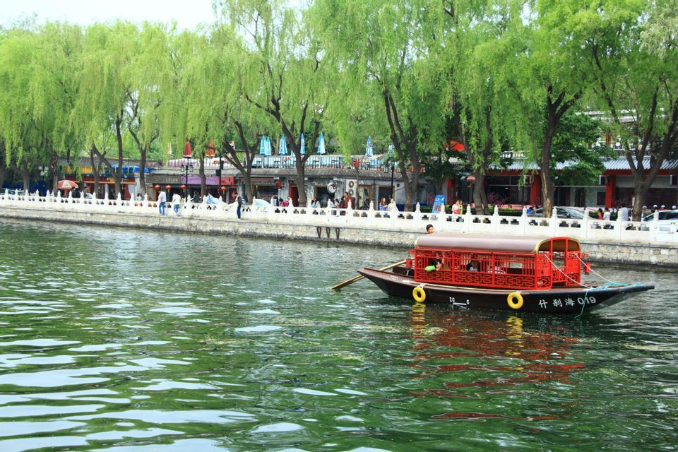Un bateau rouge traditionnel, couvert, avec des passagers, vogue sur un canal le long d'une promenade arborée, animée par des personnes et des bâtiments.