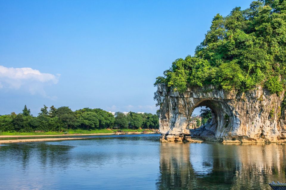 Une grande formation rocheuse couverte d'arbres avec une arche naturelle plongeant dans une large rivière sous un ciel bleu clair.