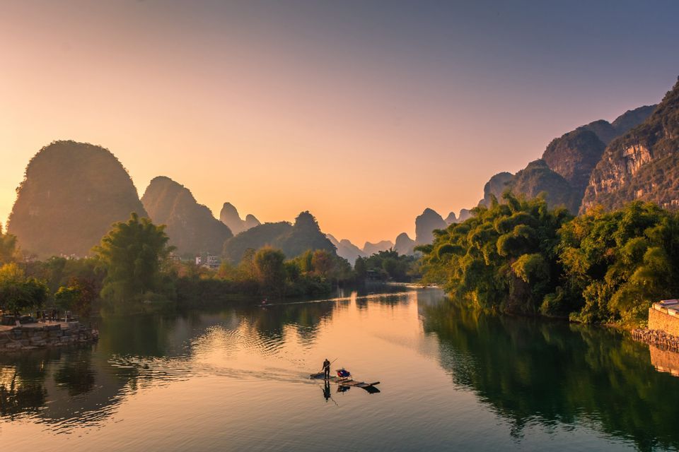 Une personne sur un radeau en bambou avance le long d'une rivière calme, entourée de montagnes karstiques et d'arbres luxuriants, au lever du soleil.