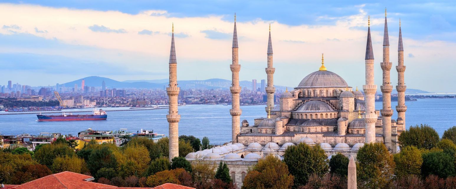 Una grande moschea con sei minareti e diverse cupole si affaccia su uno specchio d'acqua, con una nave da carico e uno skyline cittadino sullo sfondo.