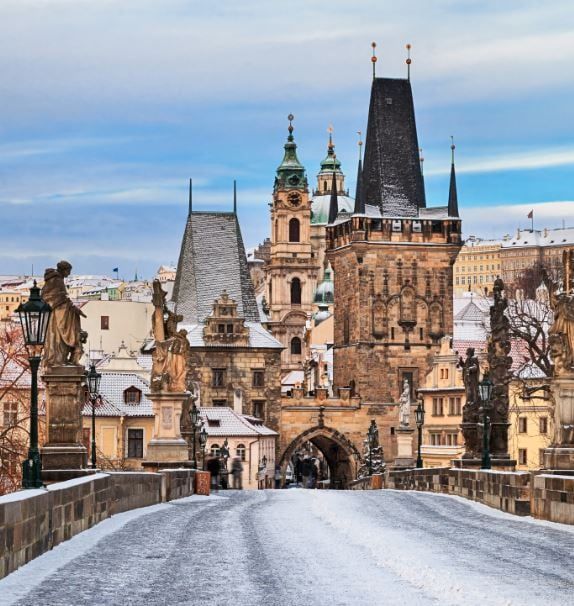 A snow-covered stone bridge lined with statues leading towards a historic tower and buildings under a blue sky.
