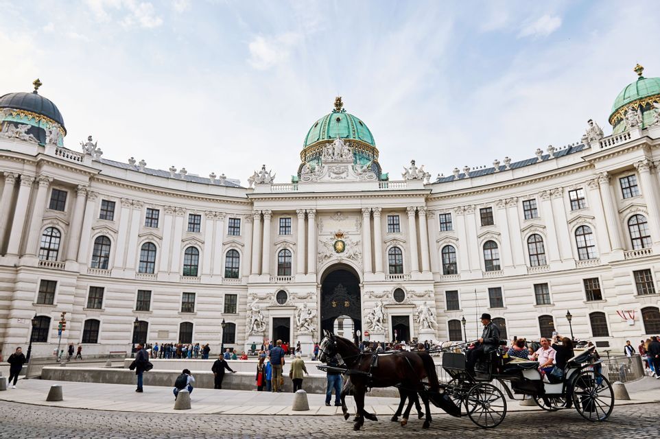 A horse-drawn carriage with tourists crosses a cobblestone square in front of an ornate white palace with green domes.
