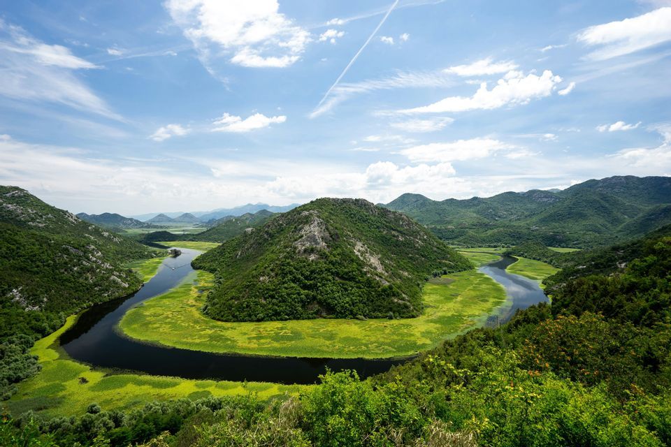 Ein breiter Fluss schlängelt sich hufeisenförmig um einen üppig grünen Hügel in einem bergigen Tal unter blauem Himmel mit weißen Wolken.