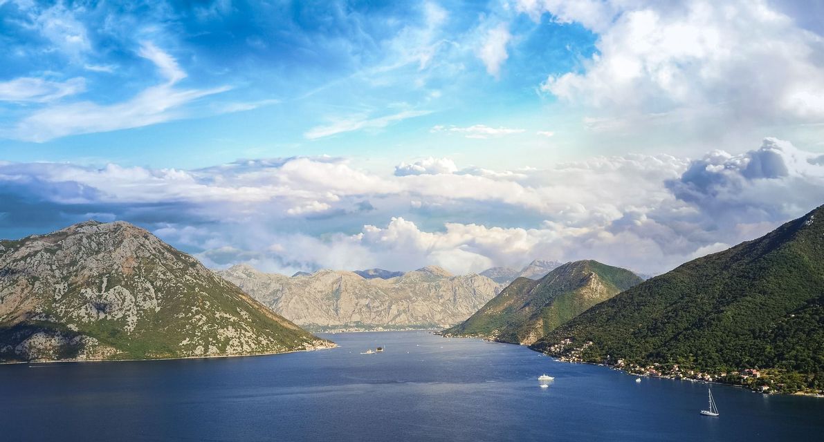 Eine Luftaufnahme einer großen Bucht, umgeben von grünen und felsigen Bergen, mit Booten auf dem Wasser unter einem bewölkten Himmel.