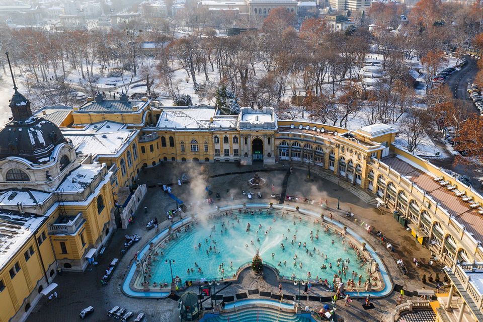 An aerial view of people bathing in a steaming outdoor pool, with snow covering the surrounding grand building and trees.