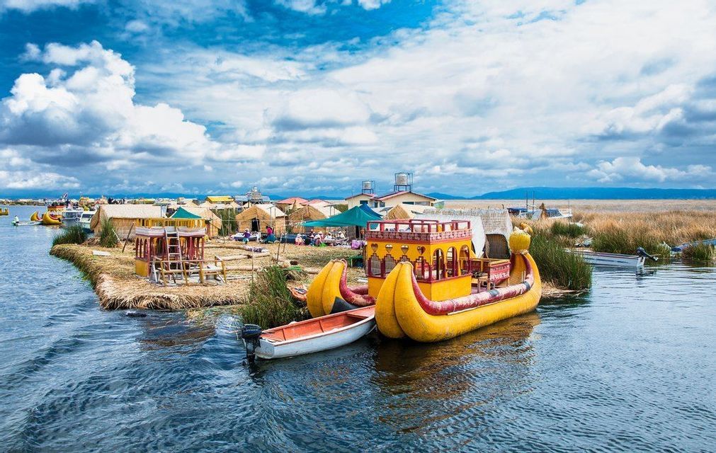 A colorful, traditional reed boat is docked at a floating island village with thatched huts on a lake under a cloudy blue sky.