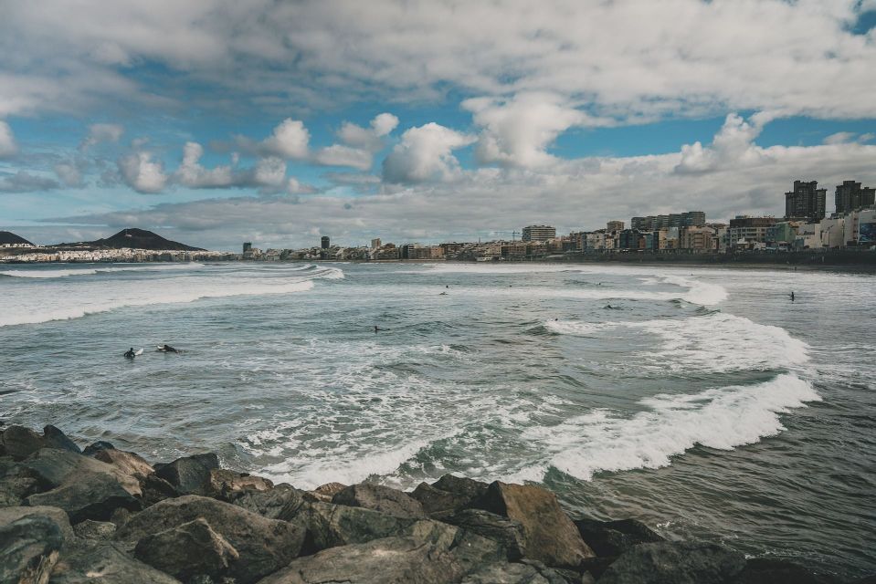 View from a rocky shoreline of surfers in the ocean with waves, a city skyline in the background, and a cloudy sky.