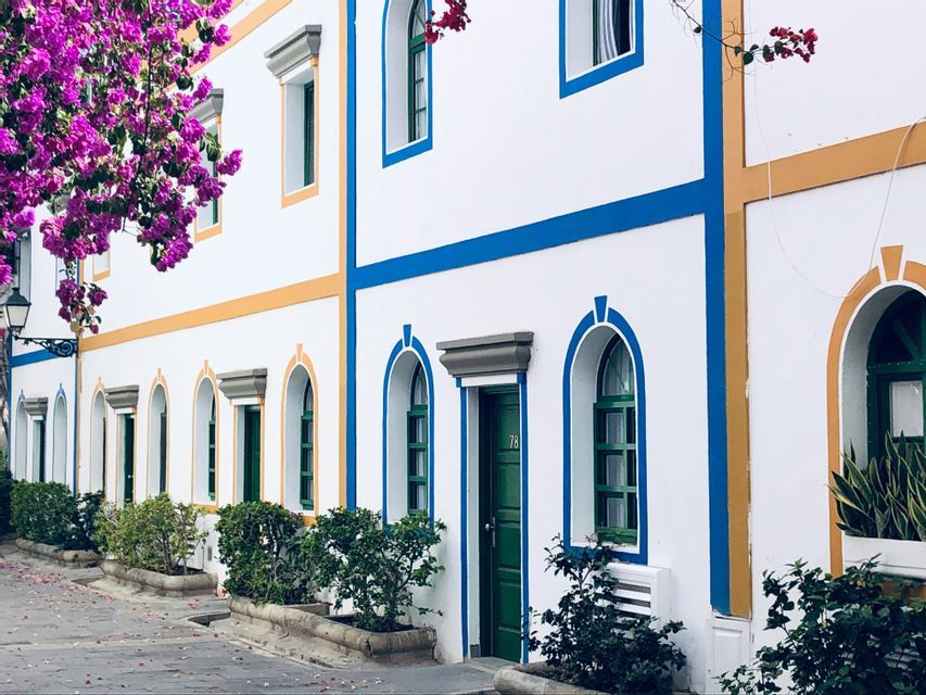 White buildings with colorful blue and yellow trim, arched windows, and green doors line a street with purple bougainvillea flowers.