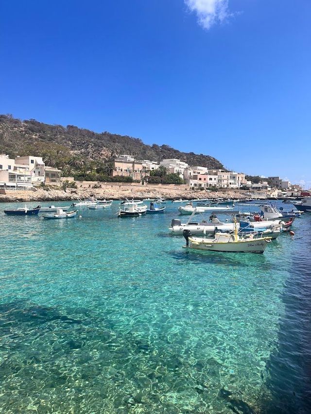 Small boats are moored in a calm bay with clear turquoise water, with a hillside town visible in the background under a blue sky.