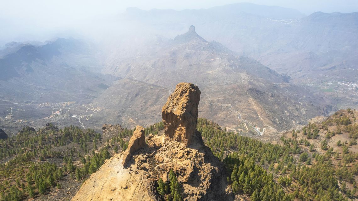 A prominent rock formation rises above a pine forest, with a vast, hazy mountain landscape in the background.